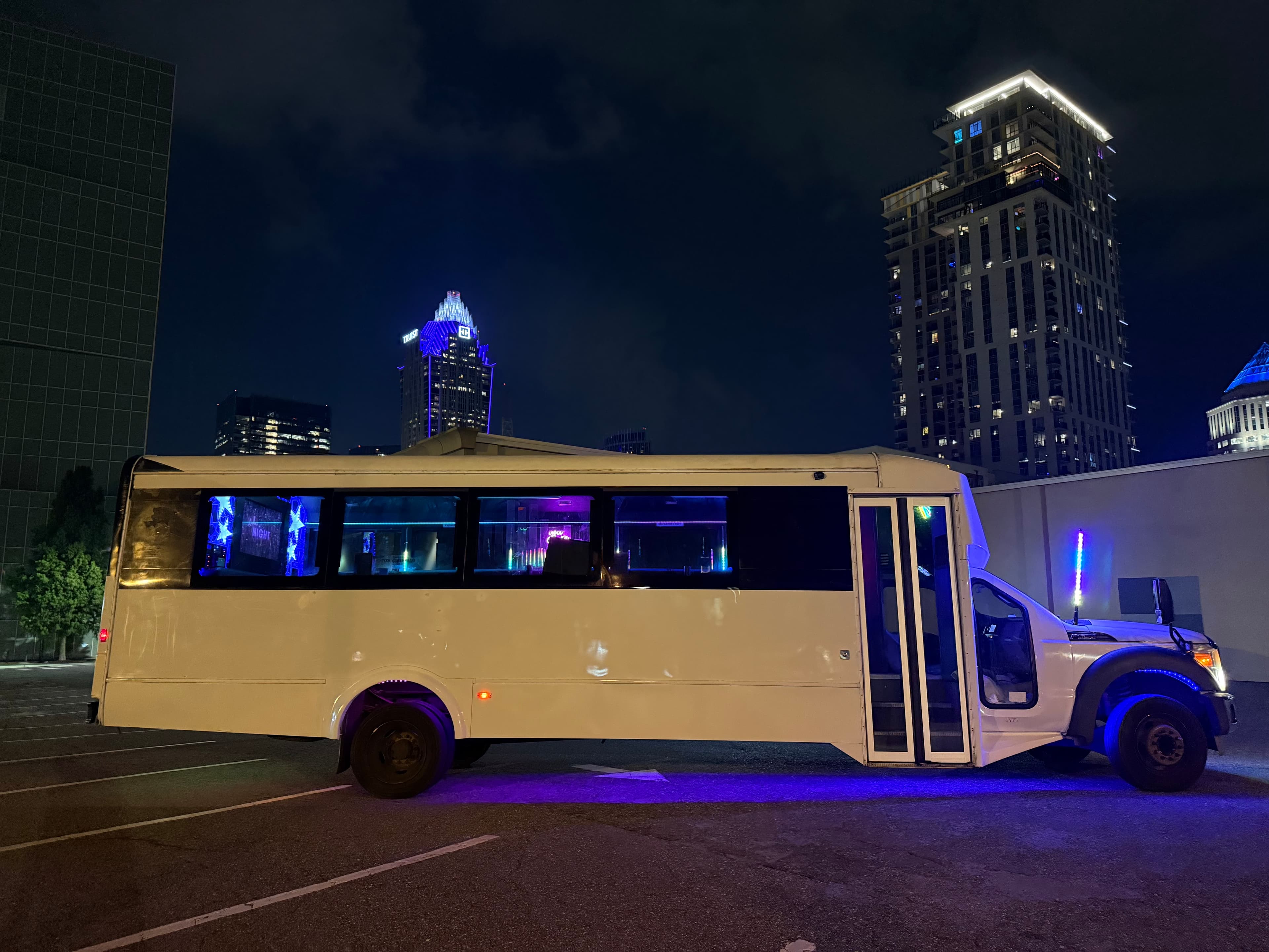 White Charlotte party bus side profile at night with blue underglow and city skyline